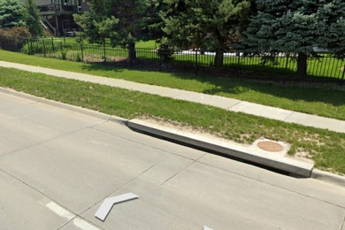 Street view of a storm drain built into the curb of a road with a pedestrian path and trees in the distance.